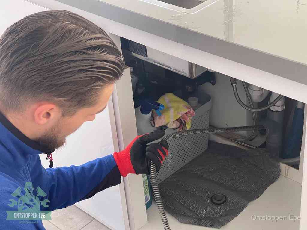 Een monteur voert een flexibele ontstoppingsveer in onder het keukenkastje, langs de sifon, om de gootsteen te ontstoppen in Epe
