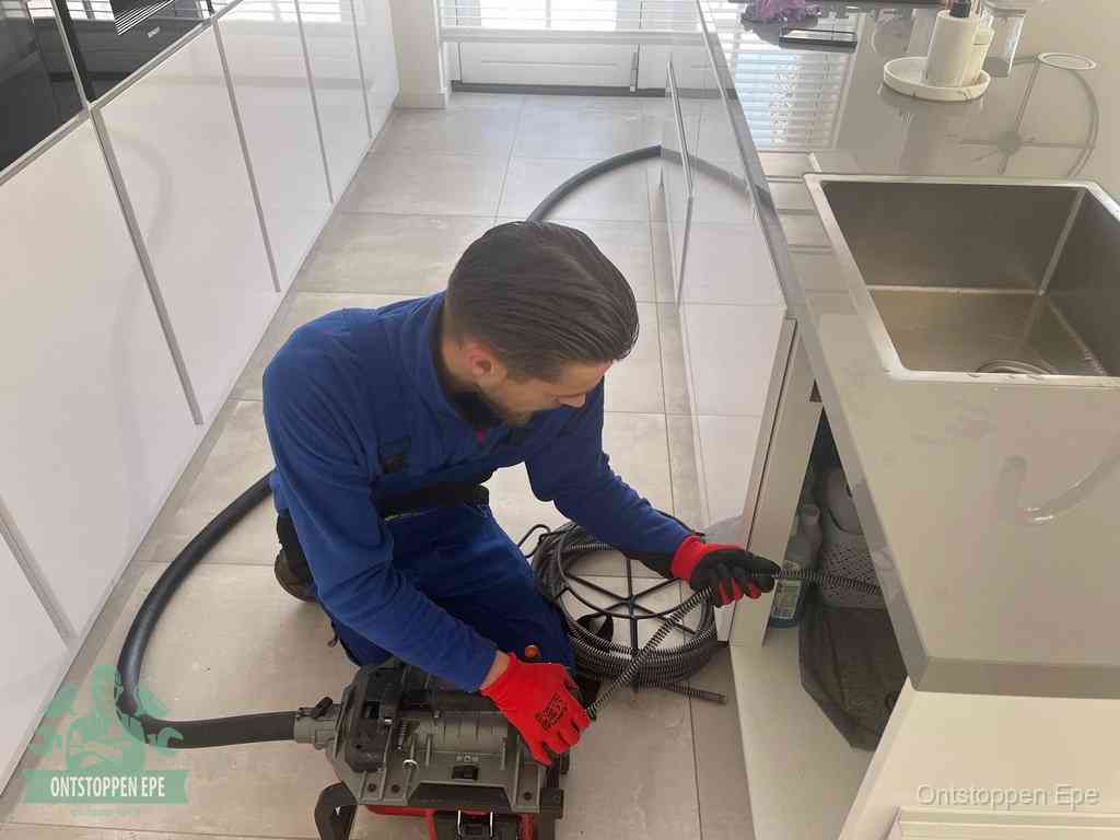 Een monteur ontstopt de keukenafvoer onder de spoelbak met een elektrische veermachine in Epe