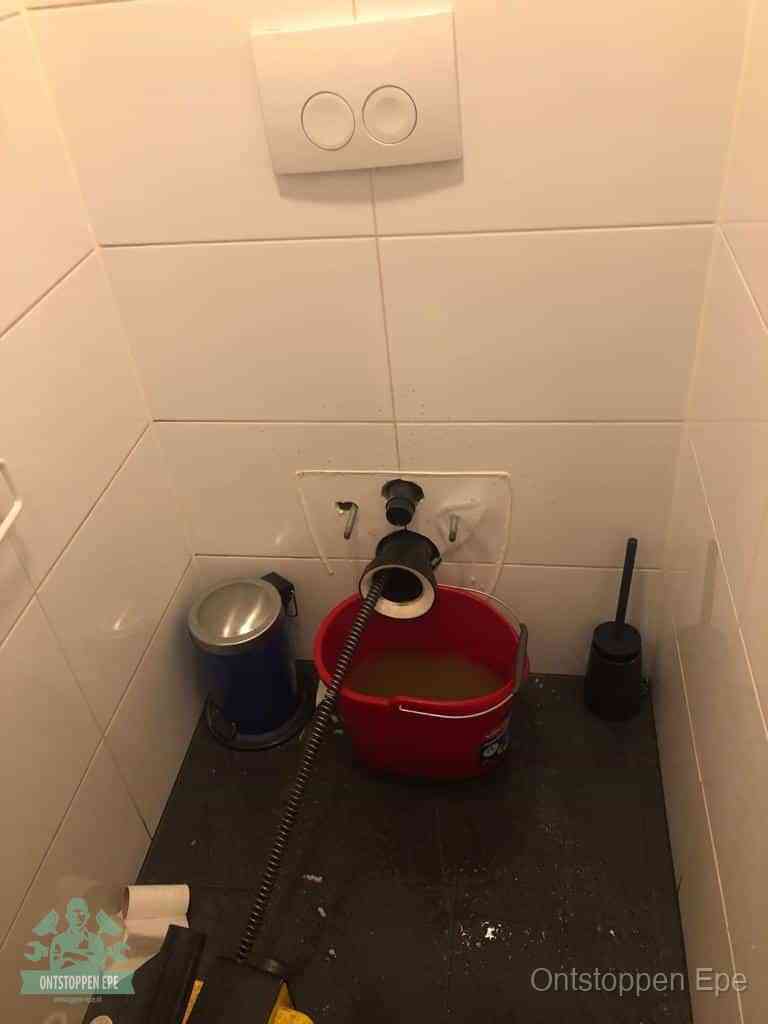 Een ontstopte wand-wc met verwijderde toiletpot waar een ontstoppingsveer in de wanduitgang verdwijnt, het achterhalen waar de verstopping zit in Epe