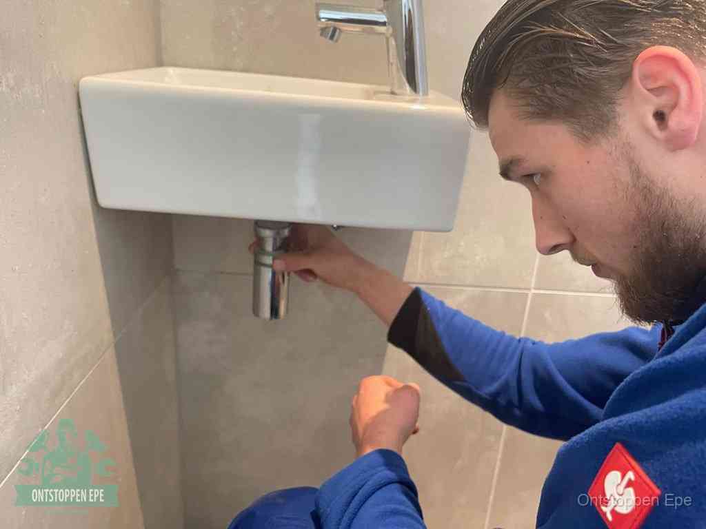 Een monteur draait met de hand de zwanenhalssifon onder een fonteinwasbak in de toiletruimte los om de bron van rioollucht in Epe te vinden